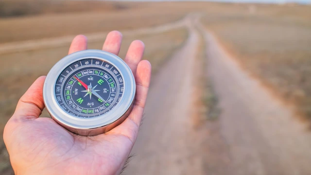 Mão aberta segurando uma bússola prateada diante de uma estrada de terra que atravessa um campo seco ao entardecer