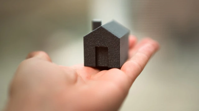 Mão aberta segurando uma pequena casa de miniatura cinza, com o fundo desfocado
