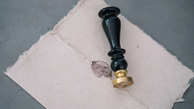 Carimbo de lacre com cabo preto e base dourada ao lado de um selo de cera já gravado, sobre papel artesanal bege