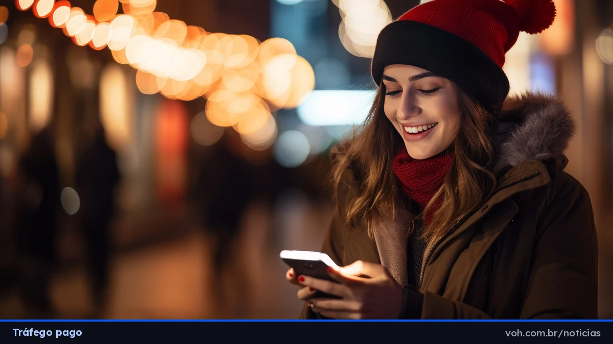 Mulher sorri olhando para o celular à noite, numa rua com luzes amarelas penduradas, usando gorro e cachecol vermelhos